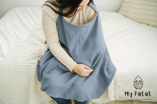 Mother sitting on white bed wearing grey muslin nursing cover while breastfeeding baby